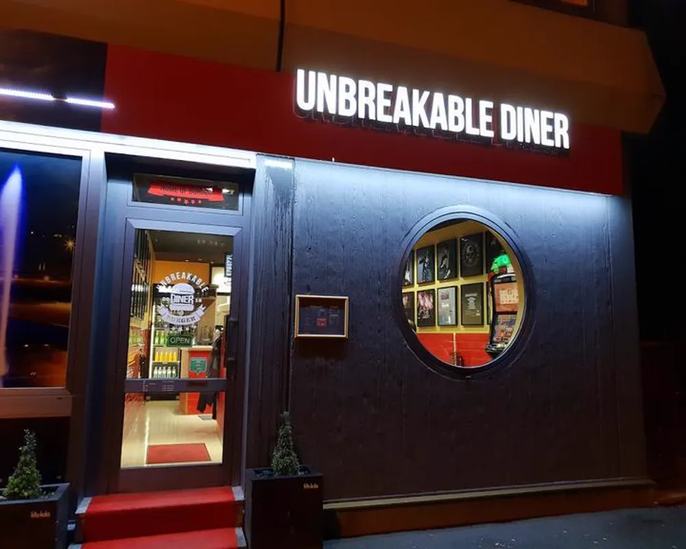 Die beleuchtete Fassade des Unbreakable Diner bei Nacht mit rotem Teppich vor der Tür